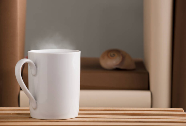 weiße Tasse Kaffee mit heißem Dampf, auf dem Schreibtisch mit Nautilus-Schale und Büchern, grauer Hintergrund. - Foto, Bild