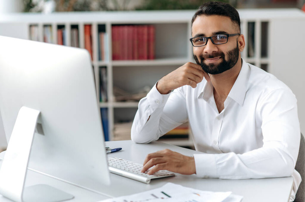 Portrait successful smart happy handsome Indian businessman, freelancer or manager with beard, wearing white shirt, sitting at table with computer in office, looking at camera and smiling friendly - 写真・画像