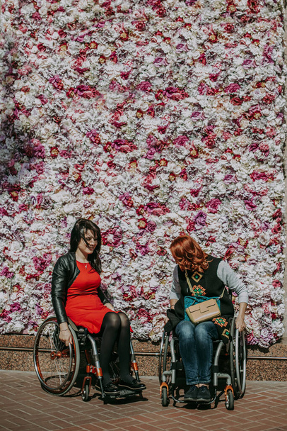 Two girls in wheelchairs against wall with artificial flowers. Walks of people with special needs in city center accessible to wheelchairs - Fotografie, Obrázek