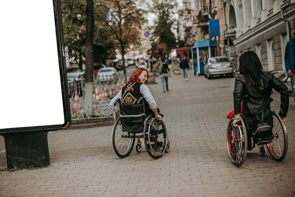 Twee meisjes in rolstoelen bij een reclamebanner met een lachend meisje. Vrouwen met speciale behoeften lopen rond Kiev in de buurt van Khreshchatyk en bewonderen de bezienswaardigheden. gelukkig meisje op een rolstoel met haar vriend - Foto, afbeelding