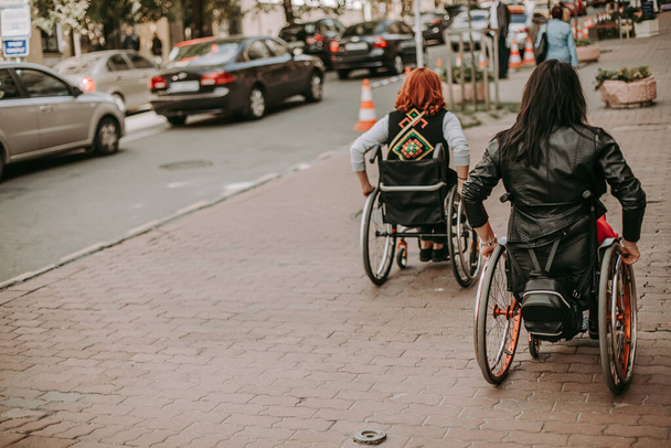 Dos chicas en sillas de ruedas cerca de un cartel publicitario con una chica sonriente. Las mujeres con necesidades especiales pasean por Kiev cerca de Khreshchatyk y admiran los lugares de interés. Chica feliz en una silla de ruedas con su amigo - Foto, imagen