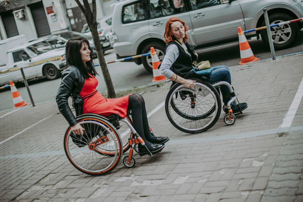 Dwie dziewczyny na wózkach jadą ulicą z samochodami w mieście. Dziewczyny podróżują na ulicę dostępną dla osób o szczególnych potrzebach na wózkach inwalidzkich - Zdjęcie, obraz
