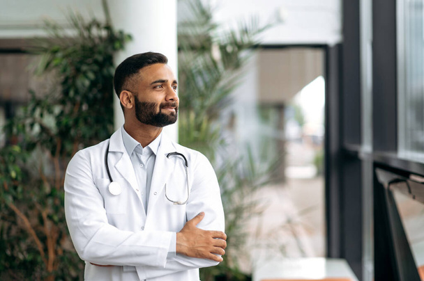 Portrait confident indian professional therapist in medical uniform and stethoscope with beard standing in hospital on blurred background looking to side and smiling friendly, dreaming about vacation - Photo, Image