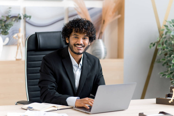 Joyful young adult successful attractive curly-haired Indian top manager, CEO or businessman in a stylish suit sits at a table in a modern office, uses a laptop, looks at the camera, smiles - 写真・画像