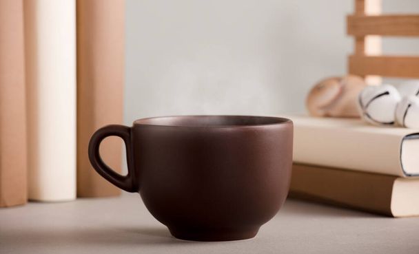 braune Tasse Kaffee mit heißem Dampf, auf dem Schreibtisch Muscheln und Bücher, grauer Hintergrund. - Foto, Bild