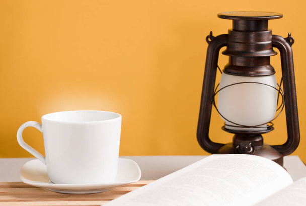 weiße Tasse Kaffee mit heißem Dampf, auf dem Schreibtisch mit offenem Buch und Sonnenlicht-Lampe, orange-gelber Hintergrund. - Foto, Bild