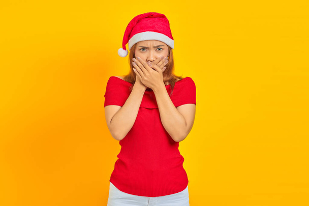 Überraschte junge Asiatin mit Weihnachtsmütze, die Mund mit Händen auf gelbem Hintergrund bedeckt - Foto, Bild