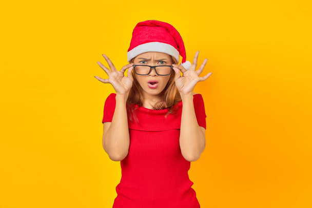 驚くべき若いですアジアの女性身に着けていますクリスマスの帽子で眼鏡をかけながらオープン口で黄色の背景 - 写真・画像