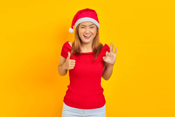 Portrait of cheerful young Asian woman showing thumbs up and ok sign gesture at camera on yellow background - Фото, зображення