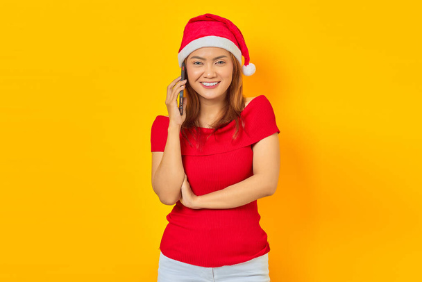 Hymyilevä nuori aasialainen nainen yllään jouluhattu puhuu älypuhelimella ja katselee kameraa keltaisella taustalla - Valokuva, kuva