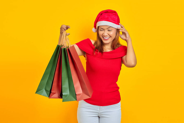 Smiling young Asian woman holding shopping bags and scratching head with hand on yellow background - Foto, Bild