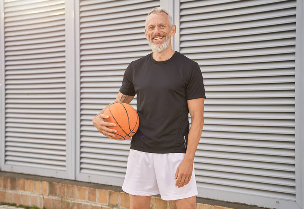 Улыбающийся мужчина средних лет в спортивной одежде держит баскетбольный мяч и смотрит в камеру, стоя на улице - Фото, изображение