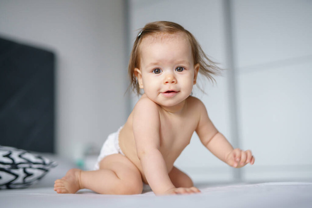 Első látásra egy kis fehér baba hét hónapos ül és mászik az ágyon otthon fényes szobában meztelenül visel pelenkát nézi a kamera boldog - Fotó, kép