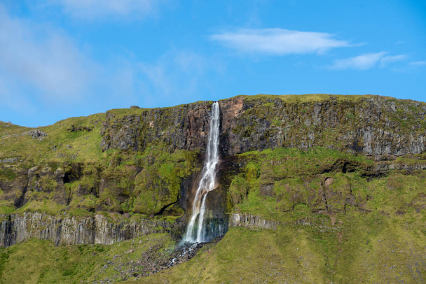 Beautiful view of a waterfall flowing down a green mountainside in Iceland. Great contrast between the blue sky and the green mountain - Foto, immagini