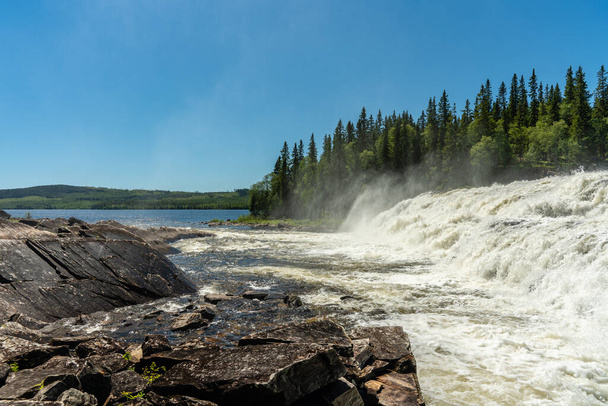 Schöne Aussicht auf einen fließenden Wasserfall in Nordschweden. Mit Kaskaden von fließendem Weißwasser und Nebel, an einem schönen Sommertag mit blauem Himmel und Sonnenschein - Foto, Bild