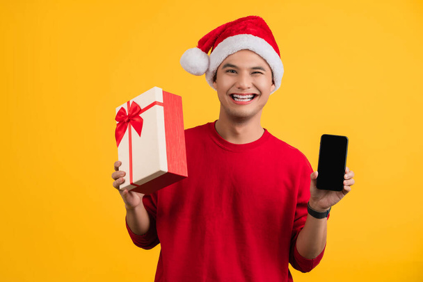 Un bel homme asiatique portant un t-shirt rouge et un chapeau du Père Noël tenant un coffret cadeau et un smartphone. Isolé sur le fond. - Photo, image