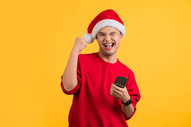 Kırmızı elbiseli ve Noel şapkalı bir adam cep telefonu ve boş ekran çalışma alanı kullanıyor. Sarı arka plan stüdyosunda izole edilmiş jest yapıyor.. - Fotoğraf, Görsel
