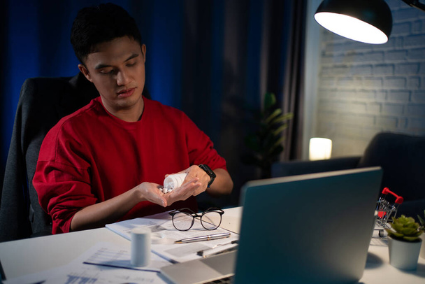 Молодой азиатский бизнесмен чувствует усталость после долгих часов работы по ночам. Он заболел и начал принимать лекарства.. - Фото, изображение