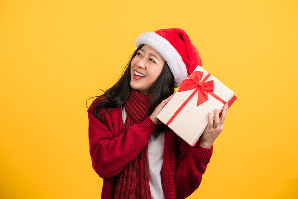 Πορτρέτο μιας ευτυχισμένης Ασιάτισσας που φοράει ένα ζεστό πουλόβερ σε ένα χριστουγεννιάτικο καπέλο στέκεται μόνη της σε ένα κίτρινο φόντο κρατώντας ένα κουτί δώρου. Κοίτα στο πλάι - Φωτογραφία, εικόνα