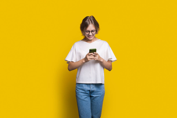 Femme blanche debout isolé sur fond jaune regardant l'écran du téléphone tout en textant un meesage. Visage souriant - Photo, image