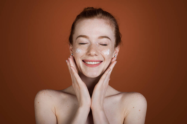 Mulher caucasiana sardenta aplicando creme cosmético em seu rosto isolado sobre fundo marrom. Mulher ruiva sorridente. - Foto, Imagem