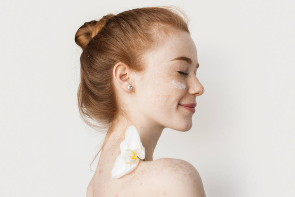 Vista laterale di una donna mezza nuda sorridente che applica la crema per il viso con un'orchidea sulla spalla isolata su sfondo bianco. Donna rossa caucasica. Cura della pelle - Foto, immagini