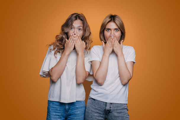 Portret dwóch zszokowanych białych kobiet zakrywających usta na pomarańczowym tle. Zaskoczeni przyjaciele. - Zdjęcie, obraz