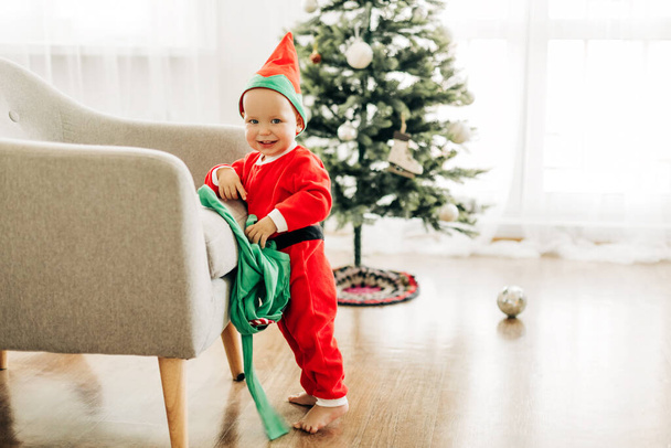 Un niño pequeño con un traje rojo está cerca de una silla sobre el fondo de un árbol de Navidad decorado. El concepto de Navidad y Año Nuevo - Foto, imagen