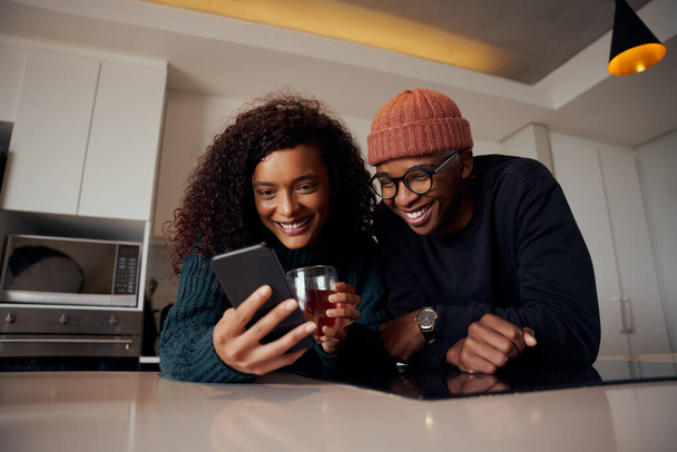 Multi-etnikai pár nevet és mosolyog telefonon, miközben a konyhában a modern lakásban - Fotó, kép