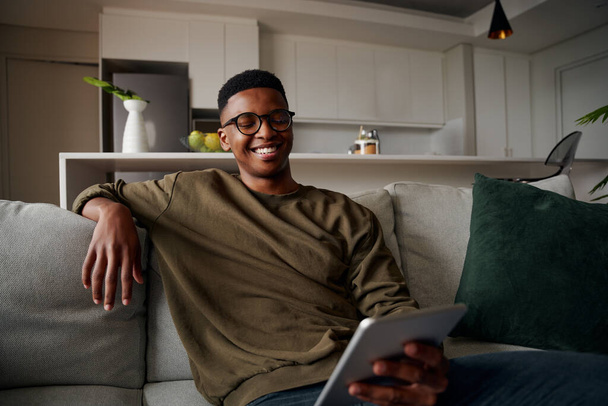 Young adult black male smiling at tablet while sitting on lounge on sofa - Photo, Image