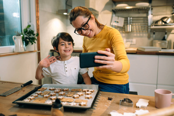 mother and child in kitchen, preparing cookies. taking selfie picture with mobile phone - Fotó, kép