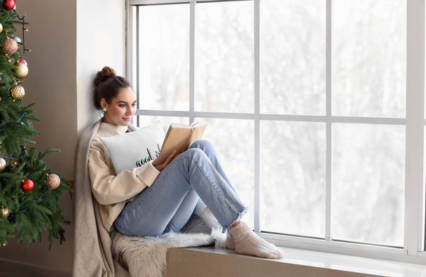Bella giovane donna con libro vicino alla finestra a casa la vigilia di Natale - Foto, immagini