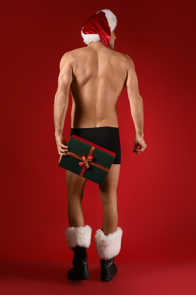 Sexy young man in Santa hat, underwear and with Christmas gift on color background, back view - Fotó, kép