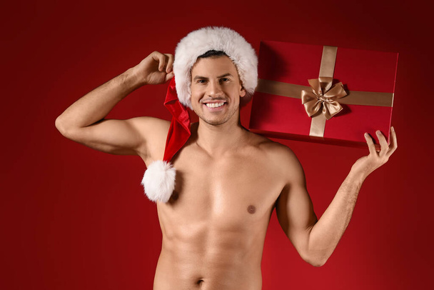 Sexy jongeman in Santa hoed en met kerstcadeau op kleur achtergrond - Foto, afbeelding