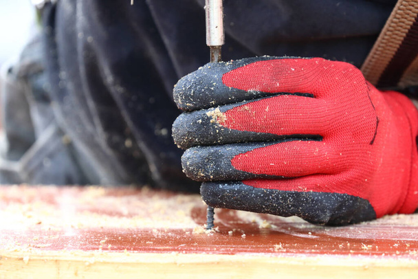 A closeup shot of a constructor's hand in a rubber glove holding a nail and drilling in a wooden piece. - Photo, image