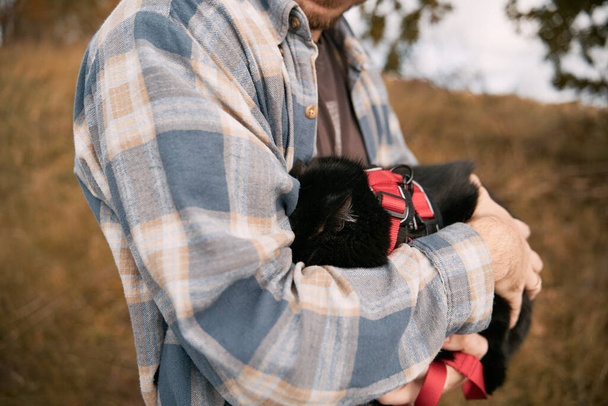 Ένας νεαρός άνδρας που φοράει καρό πουκάμισο αγκαλιάζει τη μαύρη γάτα του κατά τη διάρκεια ταξιδιού στο ύπαιθρο. Έννοια του Σαββατοκύριακου ταξιδιού στην αγροτική περιοχή με κατοικίδια ζώα στο σπίτι. - Φωτογραφία, εικόνα