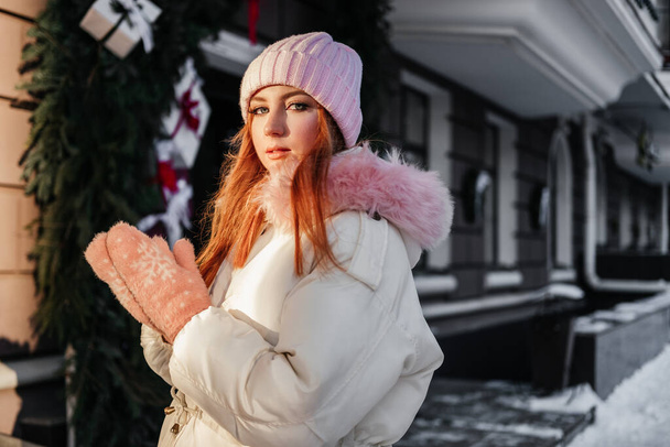Paltolu kızıl, pembe eldivenli genç bir kadın binanın önünde duruyor. Üzerinde köknar dalları ve hediye kutuları var. Kızıl saçlı kız 20 yaşında açık havada - Fotoğraf, Görsel
