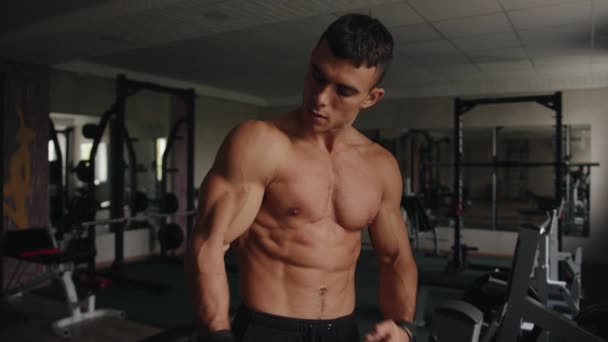 Kehonrakentaja ilman t-paitaa osoittaa pois hänen pumpataan lihaksia seistessään kuntosalilla ja osuu hänen hauiksensa nyrkillä - Materiaali, video