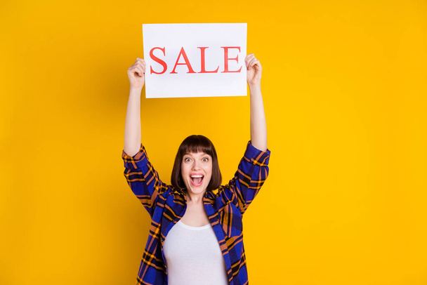 Portrait of attractive cheerful girl holding up presentation sale board ad isolated over bright yellow color background - Foto, immagini