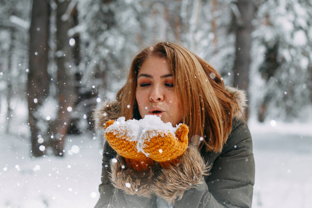A happy young girl blows on the white snow, which lies on cozy knitted woolen mustard-colored mittens. Blurred snowflakes in the foreground. Christmas holidays, Winter time. without a face. - Fotó, kép