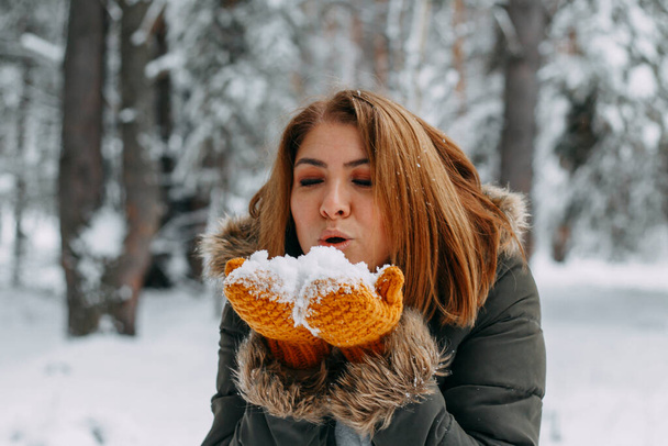 A happy young girl blows on the white snow, which lies on cozy knitted woolen mustard-colored mittens. Blurred snowflakes in the foreground. Christmas holidays, Winter time. without a face. - Фото, зображення
