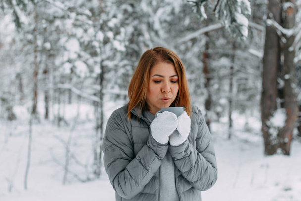 Ein schönes Mädchen in grauer Jacke und gestrickten weißen Fäustlingen trinkt ein Getränk in einem verschneiten Wald. Komfort und Wärme in der Wintersaison. Selektiver Fokus. - Foto, Bild