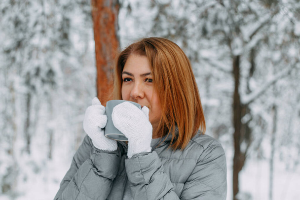 A cute girl with short hair in a gray jacket and knitted white mittens is drinking hot tea in a snowy forest. Cozy and warm in the winter season.selective focus. - Foto, imagen