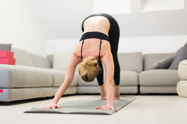 Beautiful blonde woman doing home workout indoors. Woman practice yoga at home. Fit girl using workout tutorials for healthy active lifestyle. Woman using quarantine for home workouts. - 写真・画像