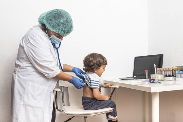 Paediatrician auscultates the back while checking the pneumonia of a blond Caucasian child in the clinic.t. checking child for infection. danger pneumonia. epidemic and pandemic covid-19. - Photo, Image