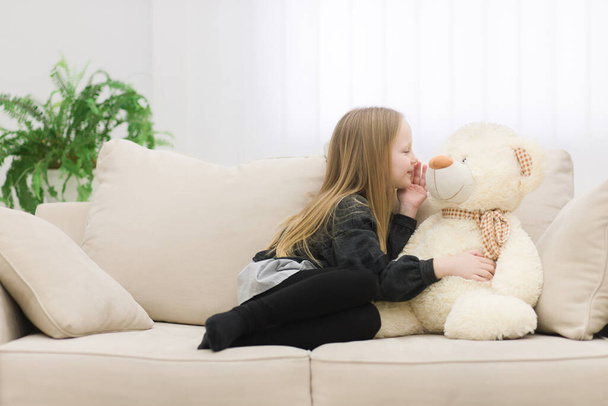 Photo of little girl sharing secrets with teddy bear on the sofa. - Fotografie, Obrázek