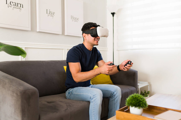 Bell'uomo latino sorridente mentre usa occhiali VR e un joystick per giocare a un videogioco di realtà virtuale a casa - Foto, immagini