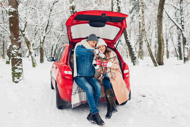 Couple in love sitting in car trunk having hot tea in snowy winter forest. Man pouring drink from thermos. People relaxing outdoors during road trip - Fotoğraf, Görsel