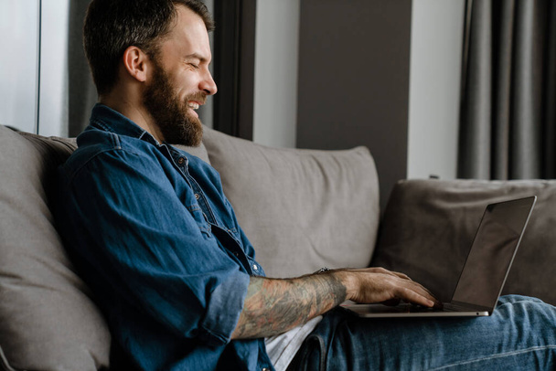 Bearded european man working with laptop while sitting on sofa at home - Fotó, kép