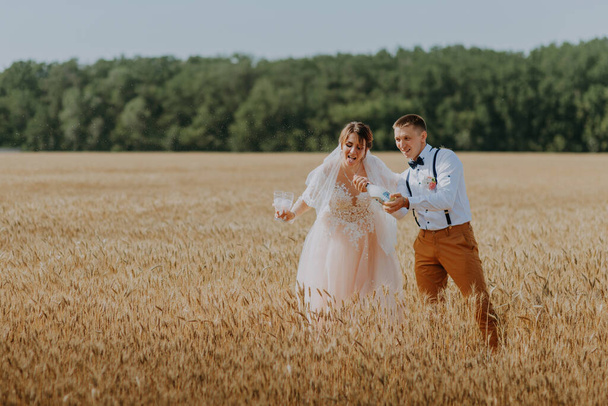 Bruid en bruidegom met champagne op de achtergrond van tarweveld. Gelukkig bruidspaar in het tarweveld. Mooie bruid in witte jurk en bruidegom hebben plezier op zomerdag. Net getrouwd. - Foto, afbeelding
